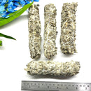 Four sage sticks tied with string, placed on a white surface with a background of blue flowers and a measuring tape for scale.