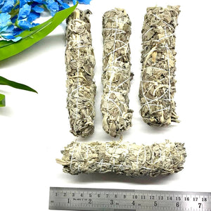 Four sage sticks tied with string, placed on a white surface with a background of blue flowers and a measuring tape for scale.